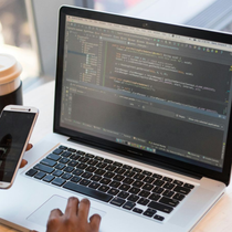 Person typing on a laptop with code on screen, holding a smartphone beside a coffee cup.