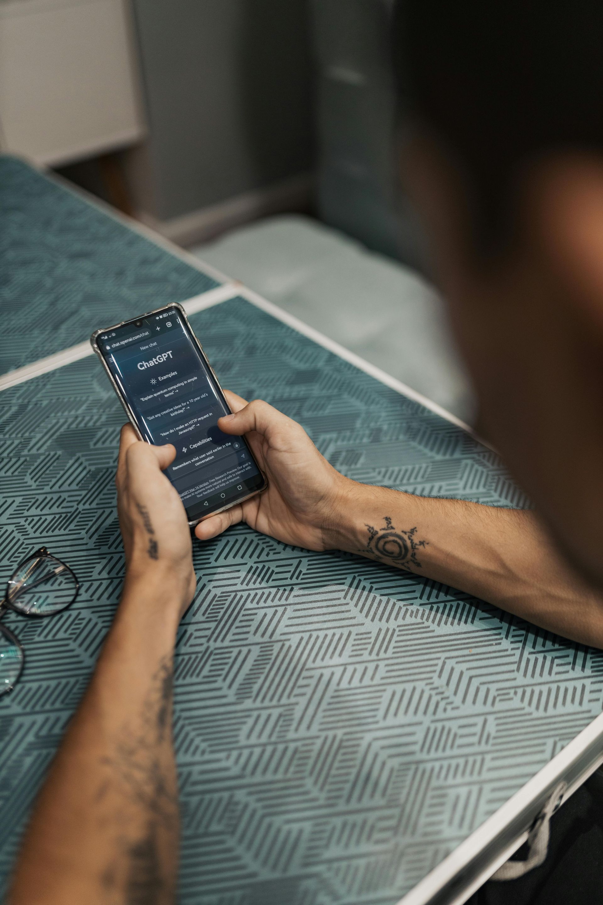 A person with arm tattoos holds a smartphone displaying a dark-themed app while sitting at a patterned teal table.