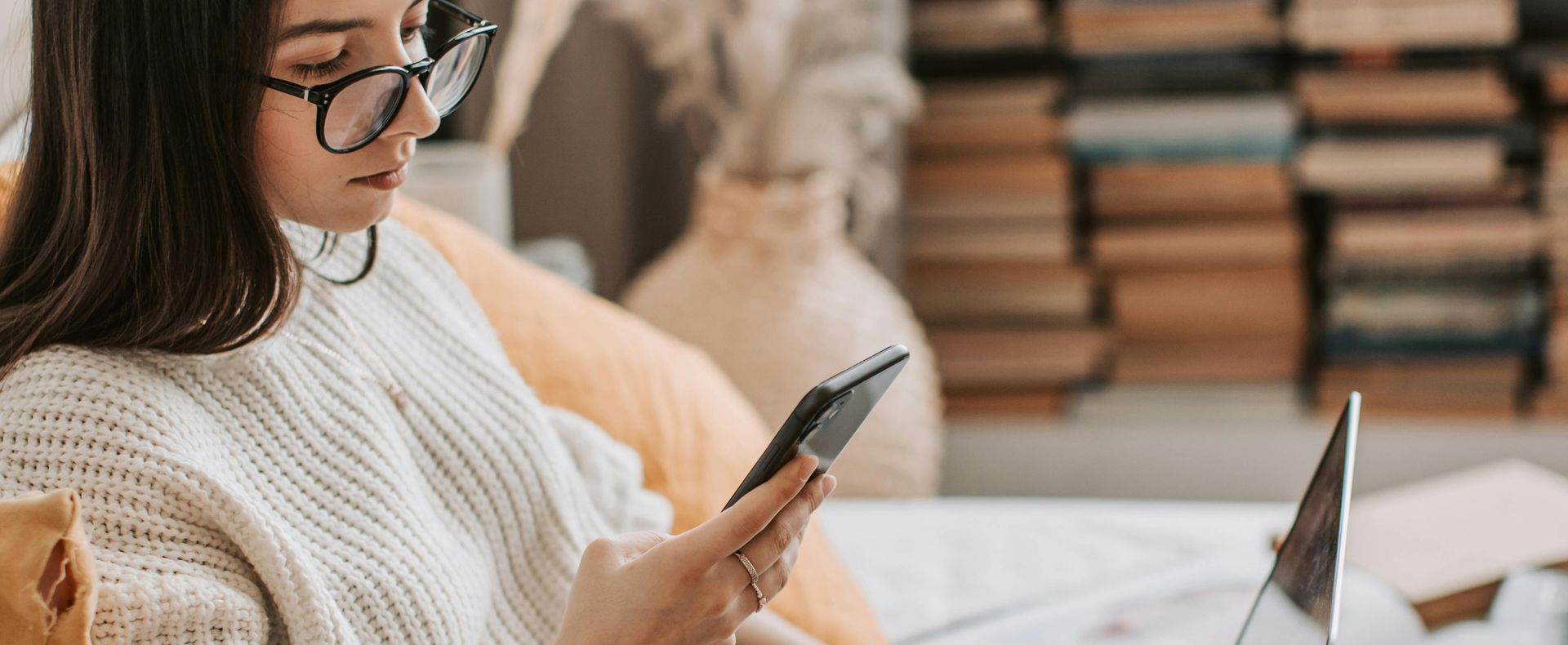 Woman with glasses looks at her phone, sitting near a laptop and a bookshelf.