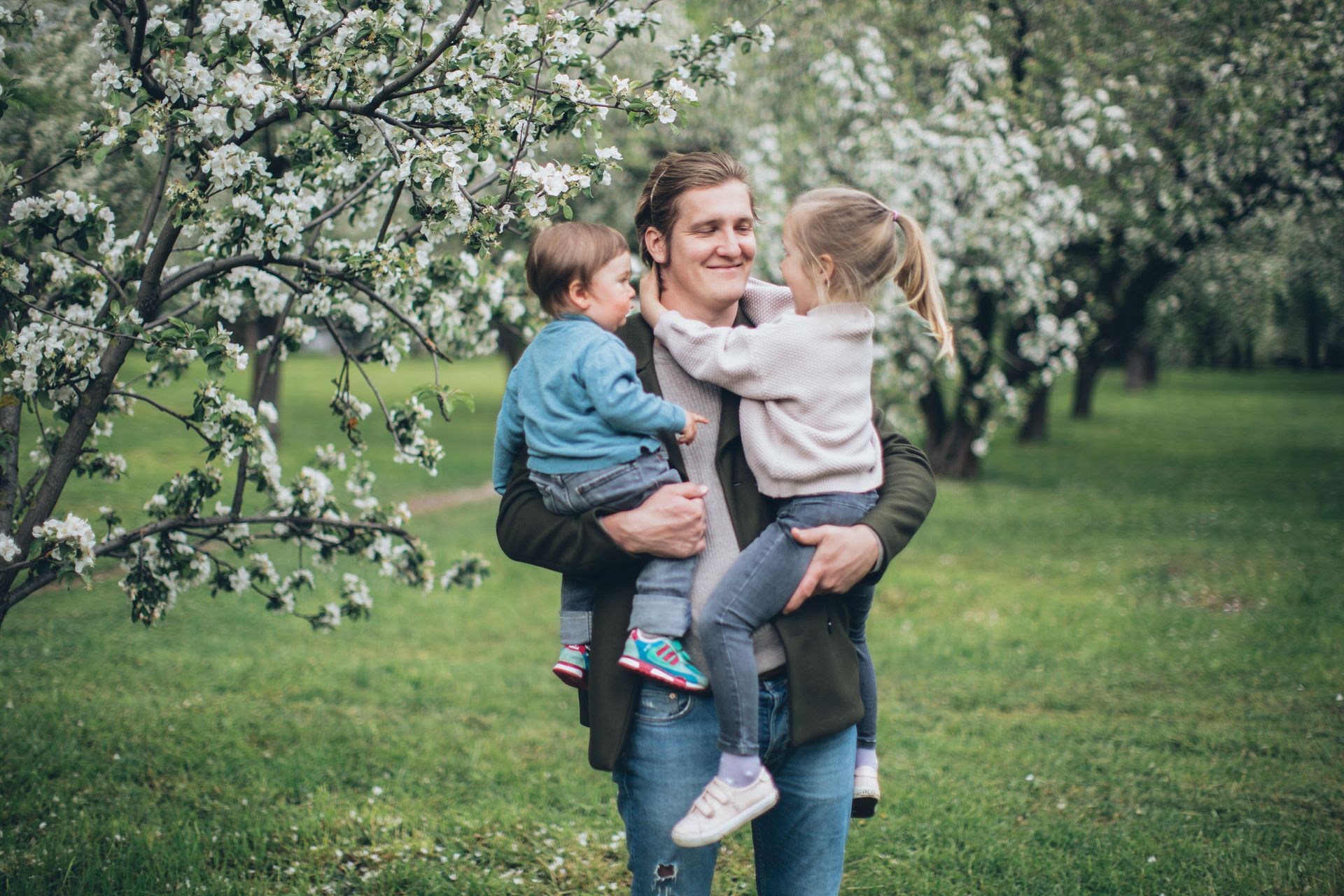 Adult holding two children in a blossoming orchard, smiling in a green field.