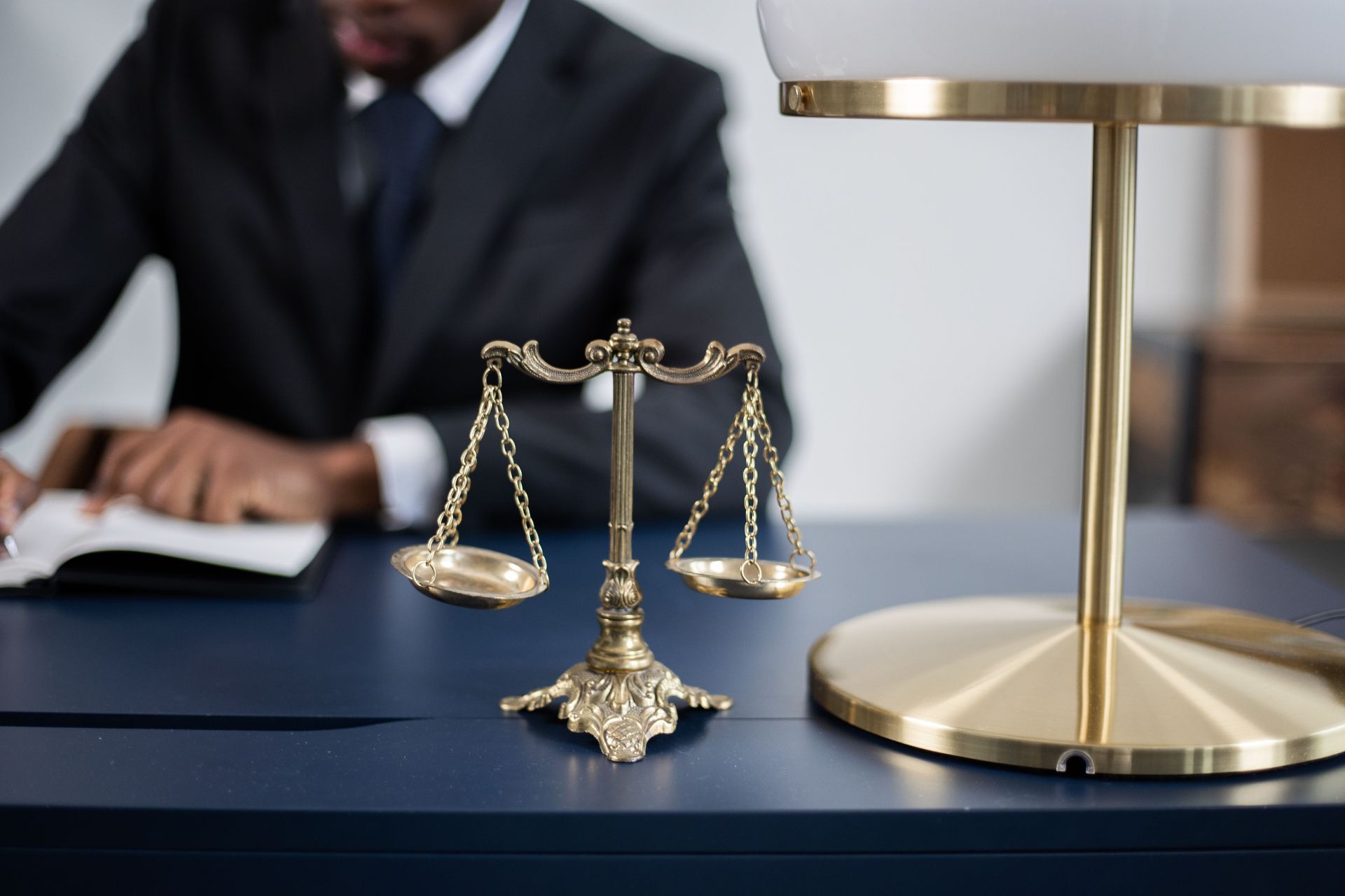 Small gold scales of justice on a desk beside a brass lamp, with a blurred professional in the background