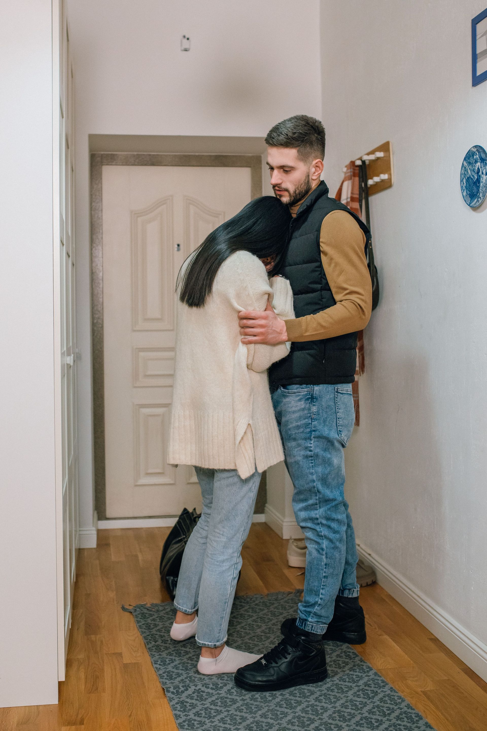 A couple embracing in a narrow hallway, one in a cream sweater and jeans, the other in a dark vest and boots.