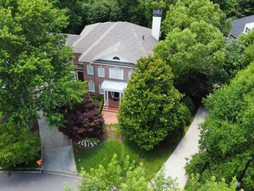 Aerial view of a brick house with a gray roof, surrounded by dense green trees and a driveway.