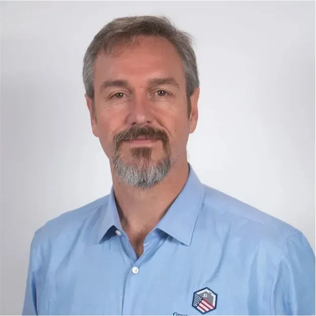 Portrait of a man in a light blue collared shirt with a small logo, standing against a white background