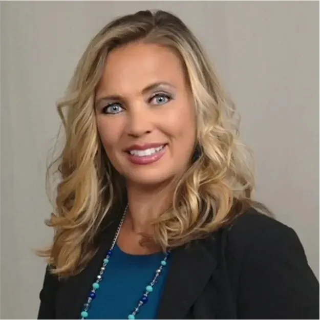 Smiling blonde woman in a black blazer and blue top against a neutral background