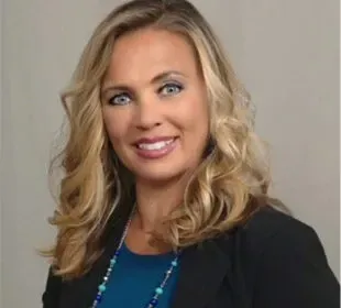Portrait of a smiling blonde woman in a black blazer and blue top against a gray background