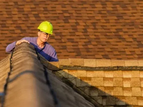 Worker in a yellow hard hat on a roof, inspecting shingles near the ridge.