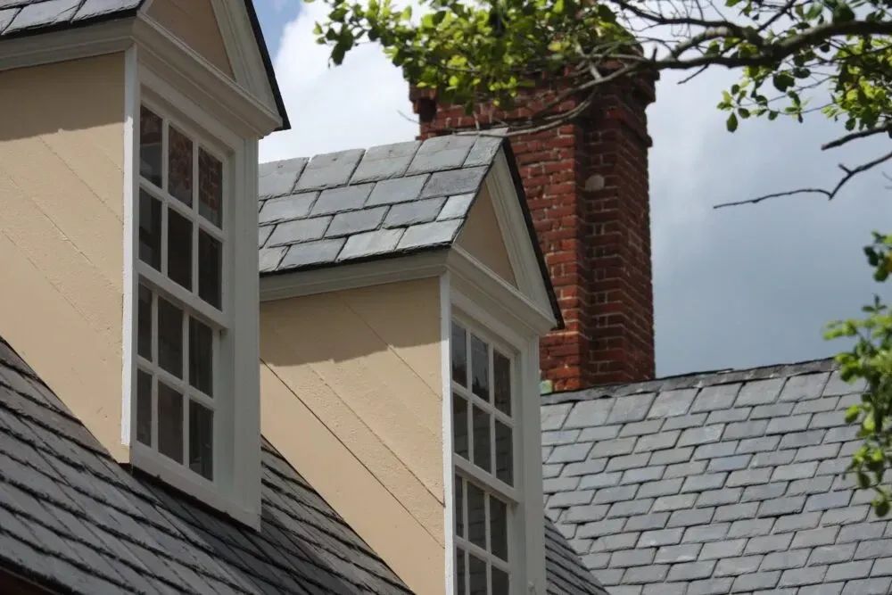 Slate-roofed beige house with dormer windows and a red brick chimney against a cloudy sky