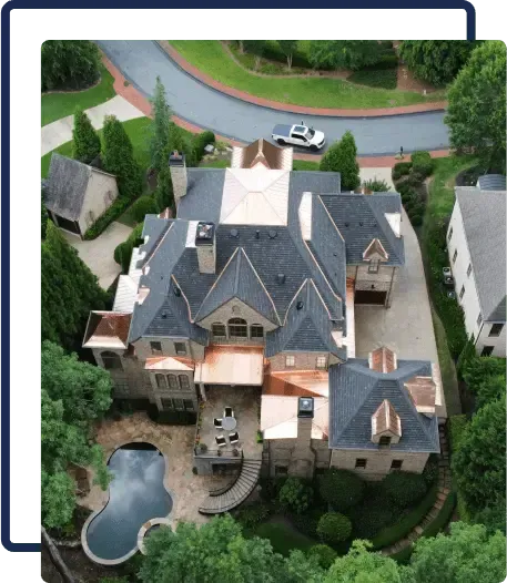 Aerial view of a large house with dark roofs, a pool, and landscaped yard near a curved road.