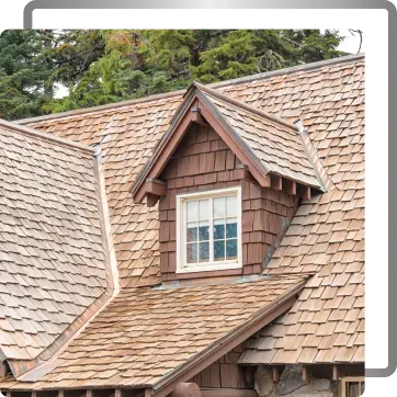 Close-up of a rustic shingled roof with a dormer window and trees in the background