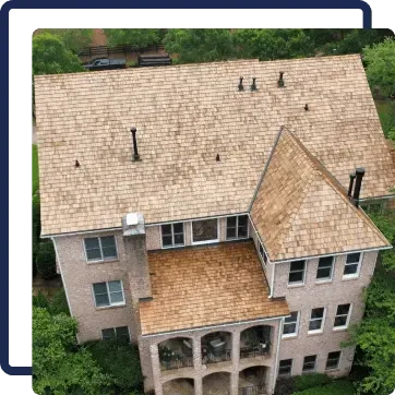 Aerial view of a beige multi-story house with a tan shingled roof surrounded by trees.