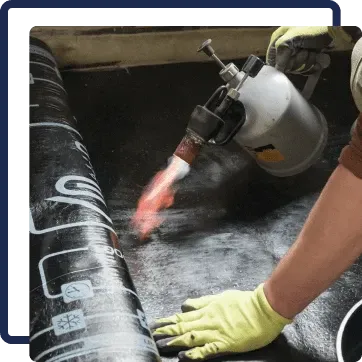 Worker using a torch to heat-weld black roofing membrane, wearing yellow gloves.