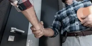 Two people shaking hands beside a door, one in a plaid shirt and one in a dark jacket.