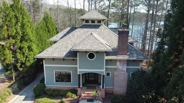 A gray house with a brick chimney and covered porch, surrounded by tall trees near a lake.
