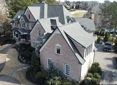 Aerial view of a large brick house with gabled roofs and a driveway in a wooded neighborhood