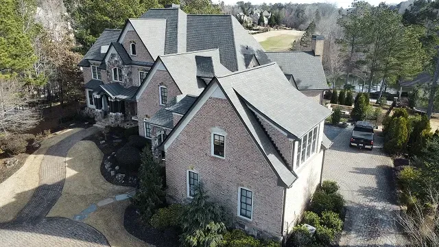 Aerial view of a large gray stone house with multiple gables and a circular driveway surrounded by trees.