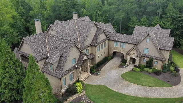 Aerial view of a large stone mansion with multiple gables, surrounded by trees and a curved driveway.