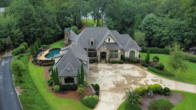 Aerial view of a large gray-roofed suburban home with a circular driveway, pool, and lush green landscaping.