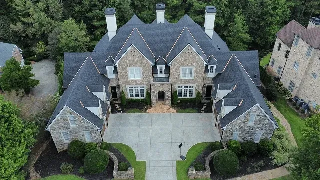 Aerial view of a large stone house with a gray roof, front driveway, and landscaped yard.
