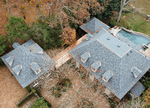 Aerial view of two gray-roofed houses connected by a walkway near trees and a pool.