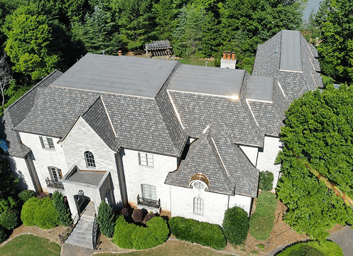 Aerial view of a large white house with gray roof surrounded by dense green trees.