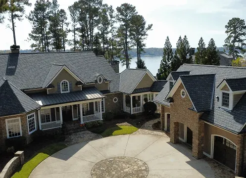 Large stone house with a dark roof and circular driveway, surrounded by tall trees near a lake.