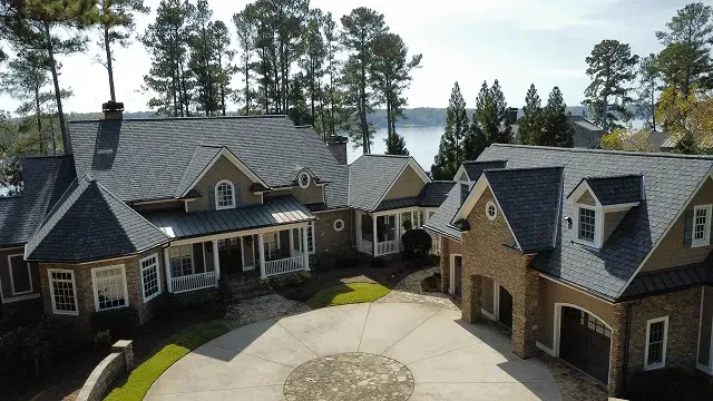 Large stone-and-shingle house with a circular driveway, surrounded by tall trees and mountains.