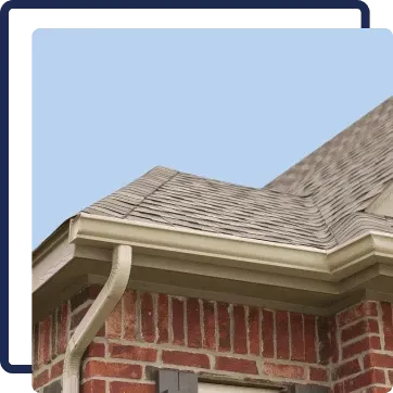Close-up of a brick house roof with light gray shingles and a white gutter against a blue sky