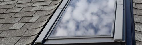 Window in a gray shingled roof reflecting clouds and sky