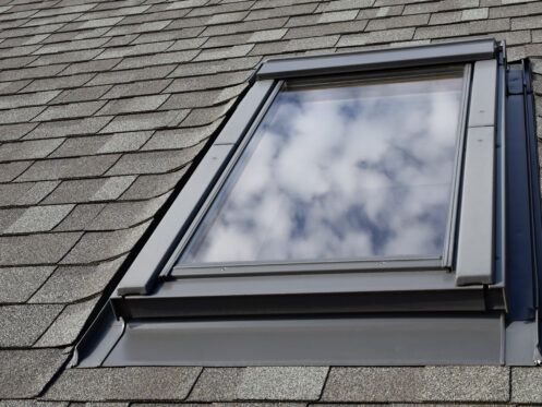 Skylight window set into a gray shingled roof, reflecting clouds in the glass