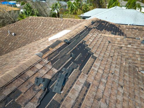 Damaged brown shingle roof with exposed underlayment and missing tiles near the ridge