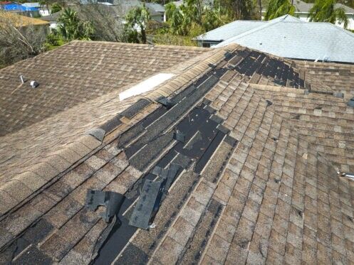 Damaged shingle roof with black tar patches and missing sections on a house