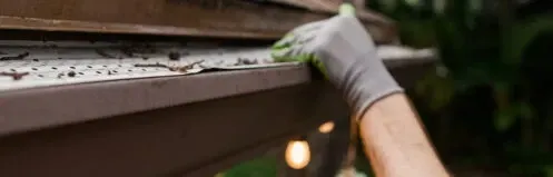 Gloved hand cleaning a roof gutter with debris near the edge of a house