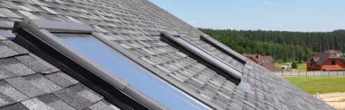 Roof with two skylight windows on gray shingles under a clear sky
