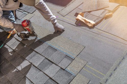 Worker installing gray roof shingles on a sloped rooftop, with tools and materials nearby.