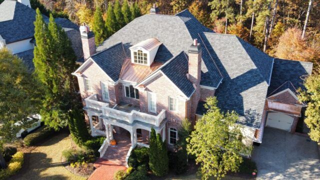Aerial view of a large brick house with gray roof, front porch, and surrounding trees.