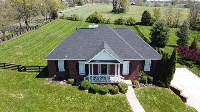 Single-story brick house with a gray roof, front porch, and green lawn in a rural setting