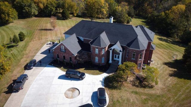 Aerial view of a large brick suburban house with a circular driveway and surrounding trees.