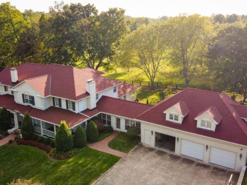 Large white house with red roof and attached garage, surrounded by trees and green lawn