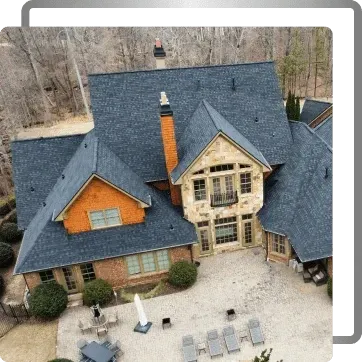 Aerial view of a large stone and wood house with a slate roof, patio chairs, and leafless trees nearby