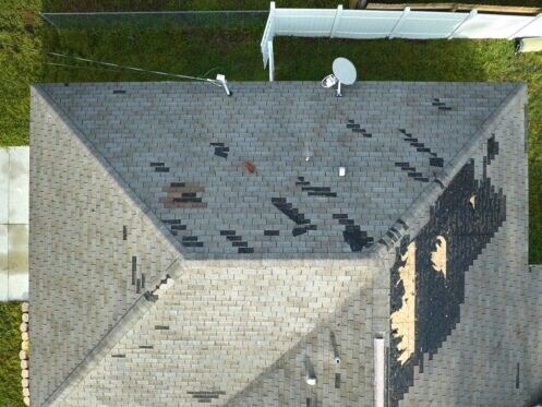 Aerial view of a gray shingled house roof with patches of damage and a satellite dish.