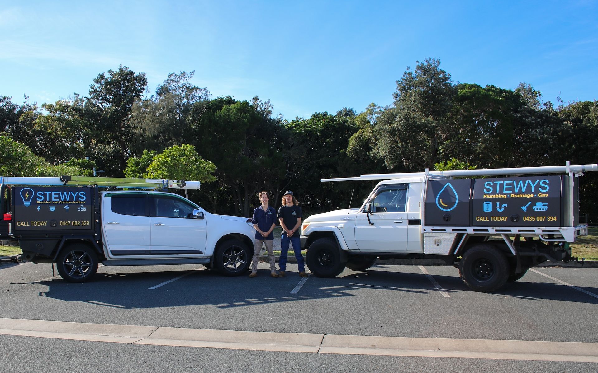 Two white utility trucks parked on a road, with two people standing between them in front of trees.