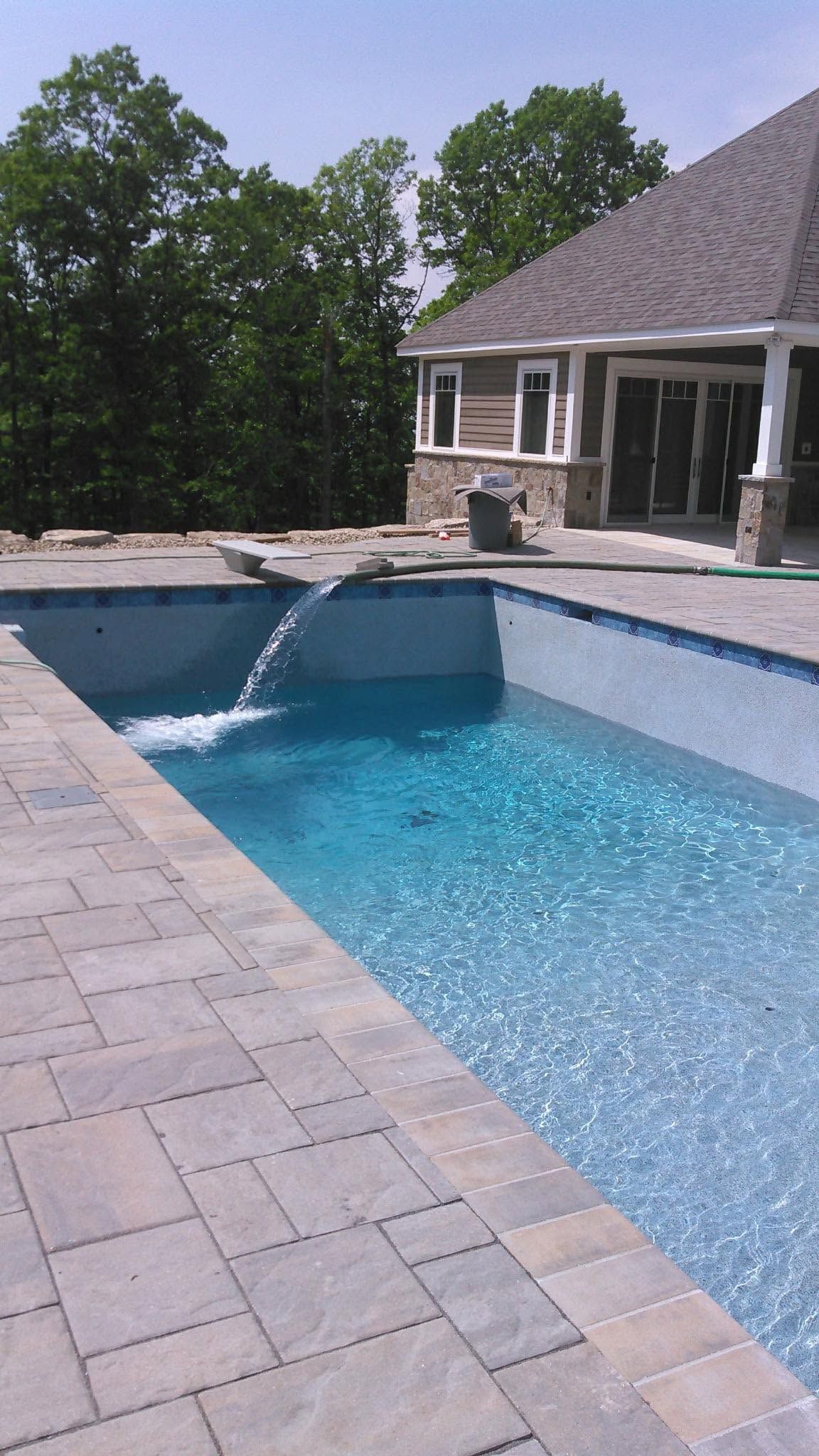 Backyard swimming pool with blue water, stone deck, and a house and trees in the background