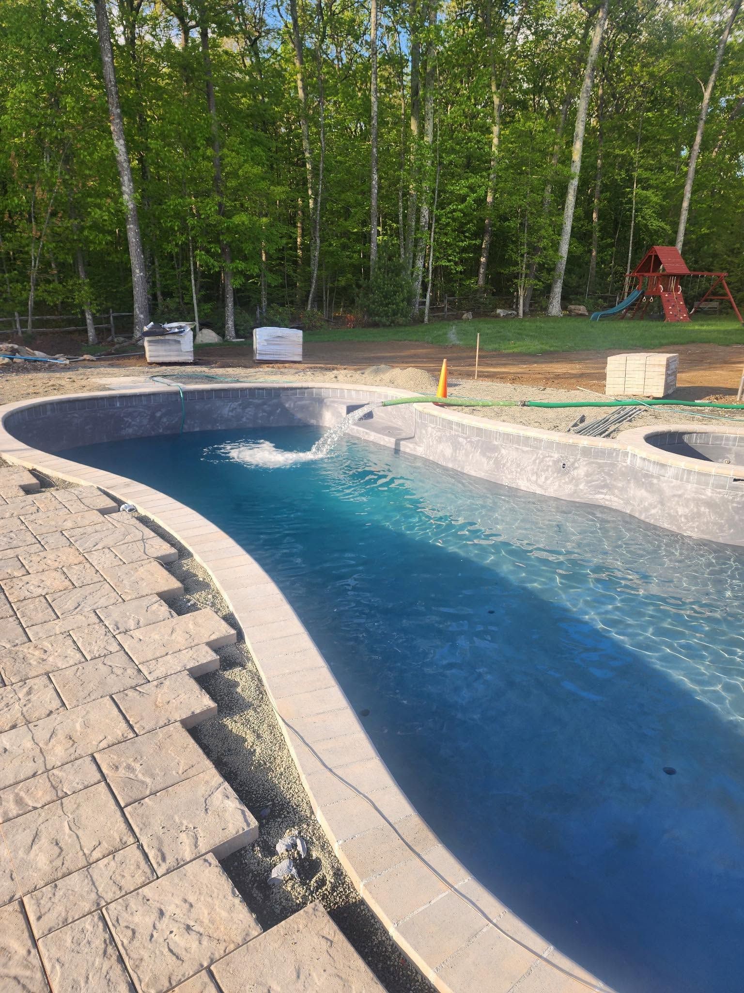 Curved backyard swimming pool with blue water, stone deck, and trees in the background