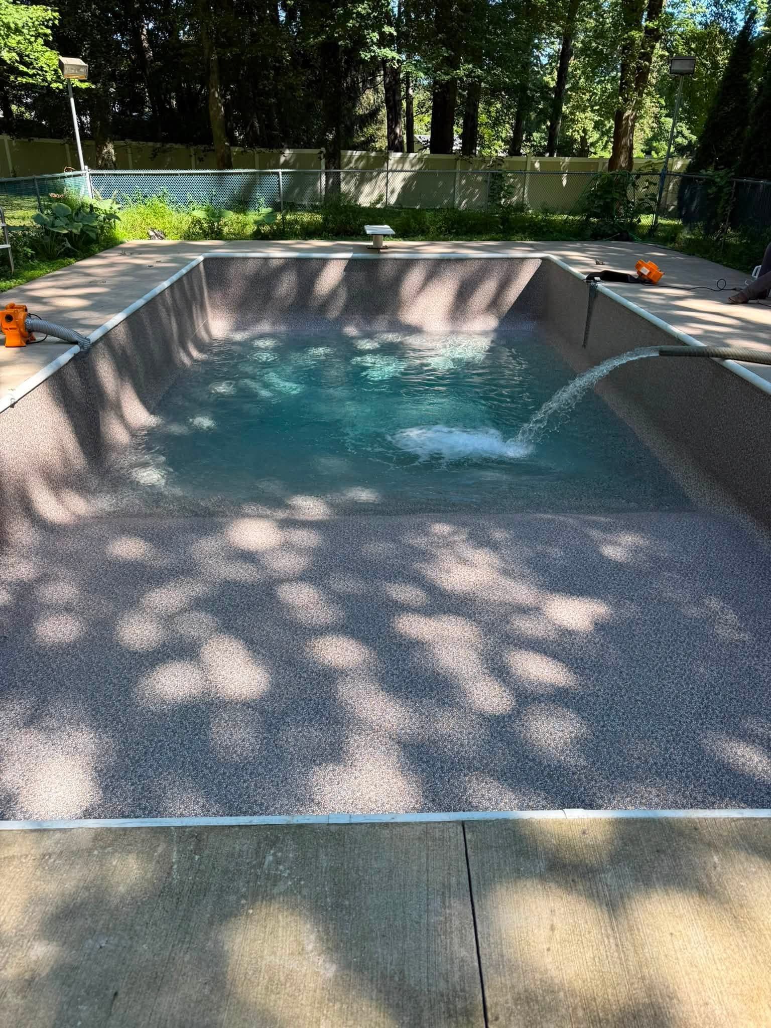 Empty backyard pool with bubbling water, surrounded by trees and dappled sunlight.