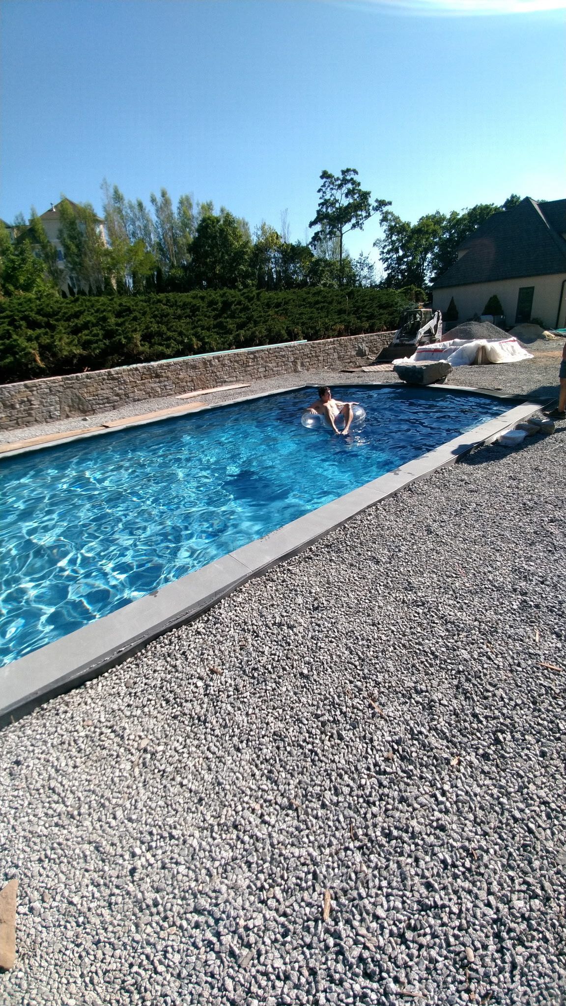 Outdoor swimming pool with two people relaxing on loungers beside a stone wall and greenery.