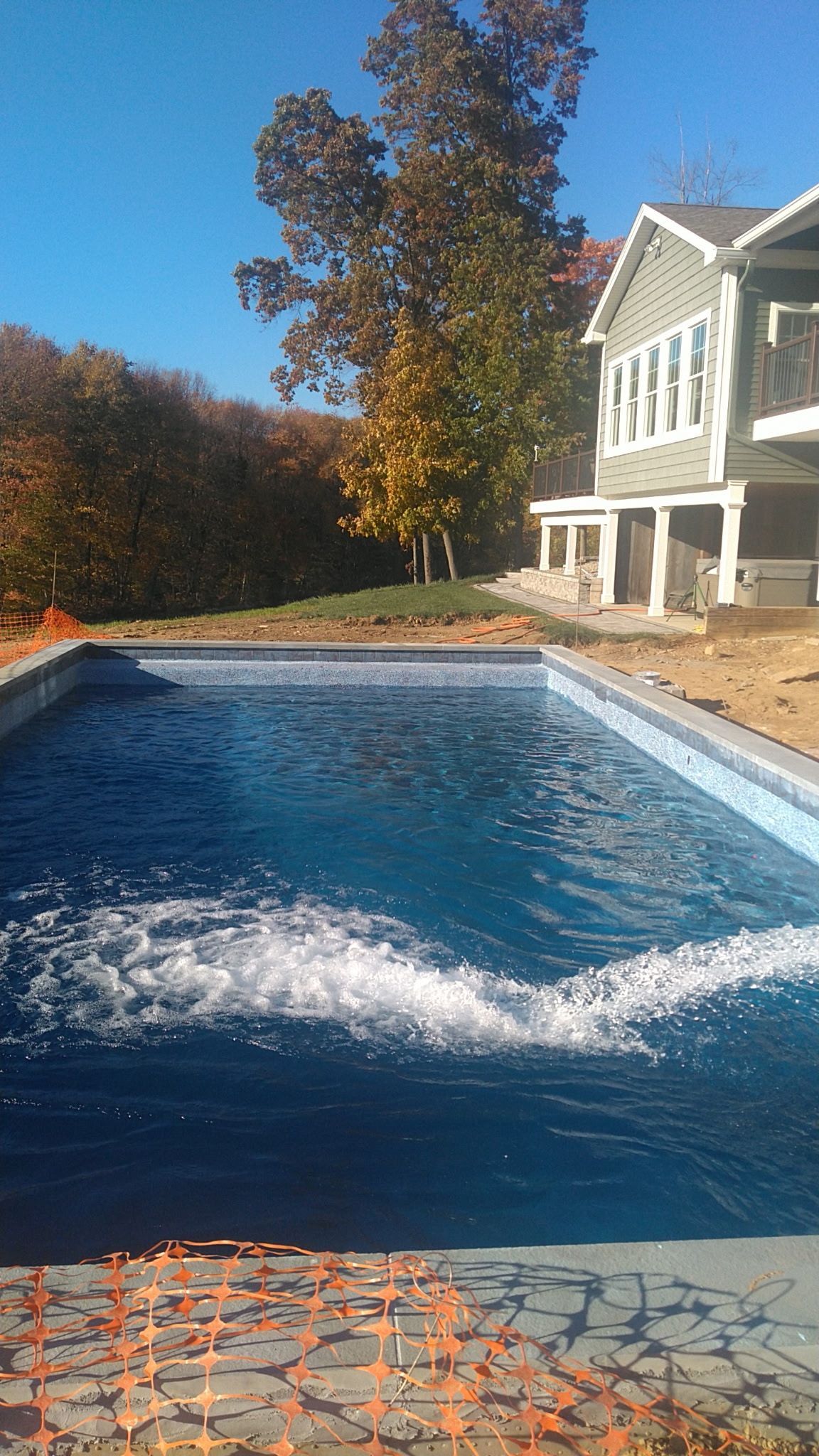 Backyard pool with blue water and a house beside autumn trees, under a clear sky
