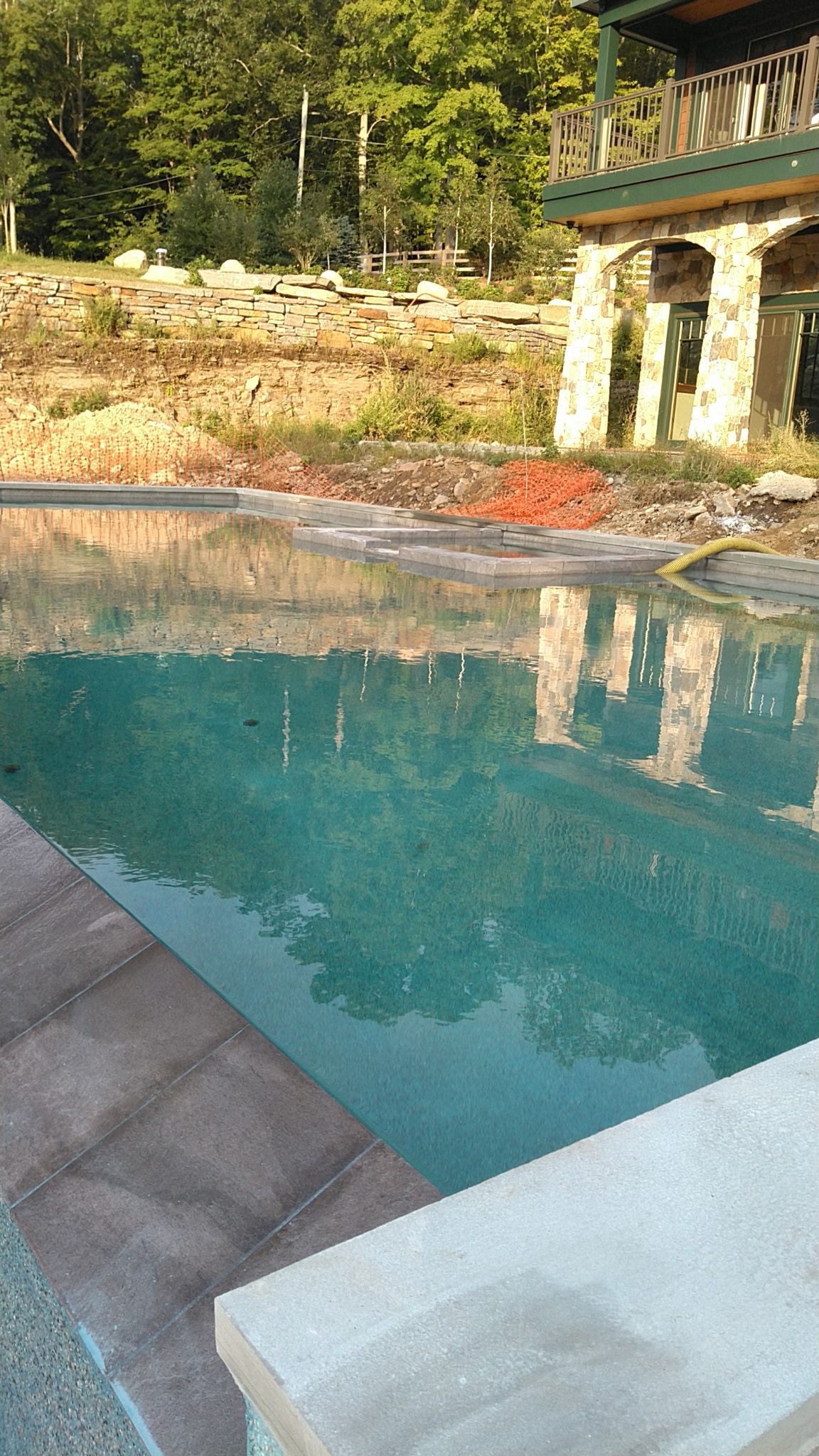 Turquoise swimming pool beside a stone deck, reflecting trees and a rustic balcony above.