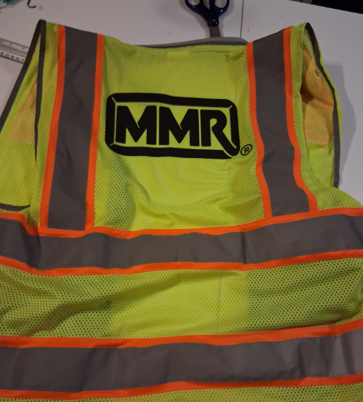 High-visibility yellow safety vest with orange reflective stripes and MMR logo hanging on a wall