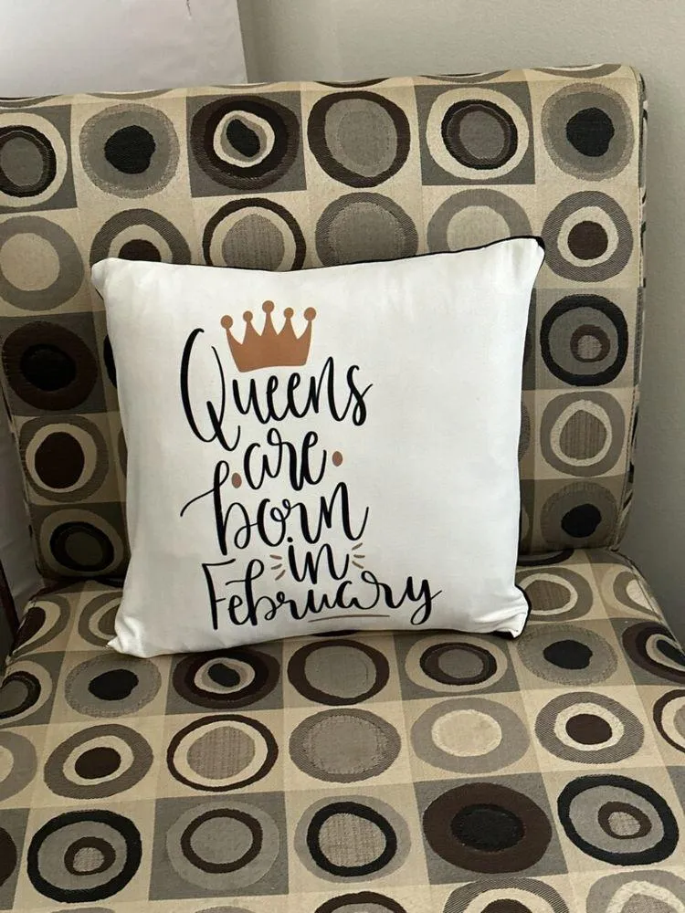 White pillow with “Queen’s have born in February” and a gold crown on a patterned couch.